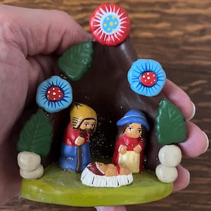 May include: A hand-painted nativity scene figurine. The figurine features a brown triangular structure with colorful floral accents. The scene includes figures of Mary, Joseph, and baby Jesus, with a green base and white accents.