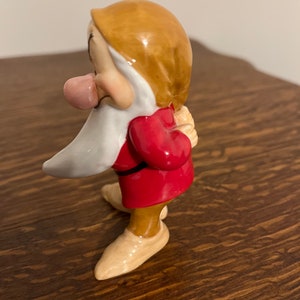 Może przedstawiać: Ceramiczna figurka krasnala z filmu Disneya Śnieżka i siedmiu krasnoludk&oacute;w. Krasnal ma na sobie czerwoną tunikę, brązowe spodnie i białą brodę. Ma wystający r&oacute;żowy język i trzyma ręce za plecami.