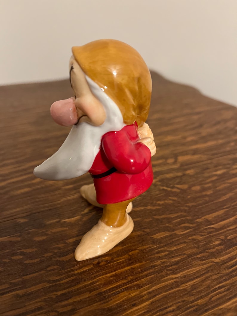 Może przedstawiać: Ceramiczna figurka krasnala z filmu Disneya Śnieżka i siedmiu krasnoludk&oacute;w. Krasnal ma na sobie czerwoną tunikę, brązowe spodnie i białą brodę. Ma wystający r&oacute;żowy język i trzyma ręce za plecami.