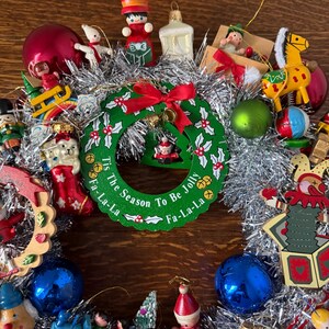 May include: A festive Christmas wreath with a green and red design, surrounded by silver tinsel. The wreath features the text "Tis The Season To Be Jolly Fa-La-La" and is adorned with various ornaments, including a red bow, bells, and miniature figurines.