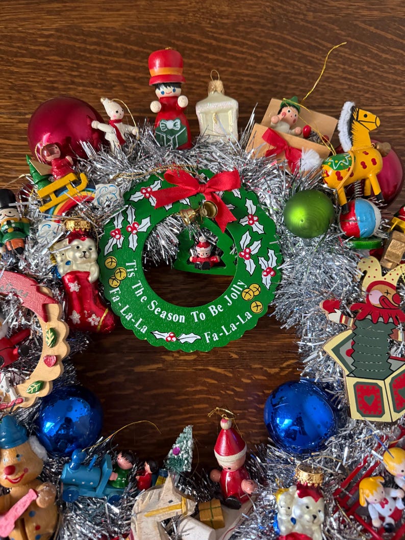 May include: A festive Christmas wreath with a green and red design, surrounded by silver tinsel. The wreath features the text "Tis The Season To Be Jolly Fa-La-La" and is adorned with various ornaments, including a red bow, bells, and miniature figurines.