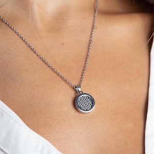 Puede incluir: Un collar de tono plateado que presenta un colgante circular con un centro texturizado y punteado. El colgante está suspendido de una cadena fina y delicada de tono plateado. El diseño es simple y metálico, mostrando un borde exterior pulido y una superficie granular detallada dentro del marco circular, creando un contraste sutil en la textura y el reflejo de la luz.