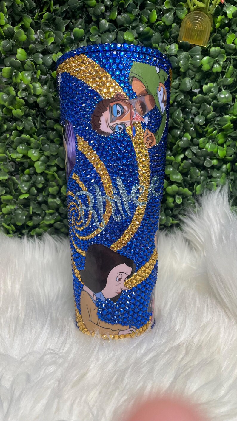 May include: A blue and gold tumbler featuring a cartoon image of a person wearing glasses and a moustache. The tumbler is covered in rhinestones and has the word "remarkable" written on it.