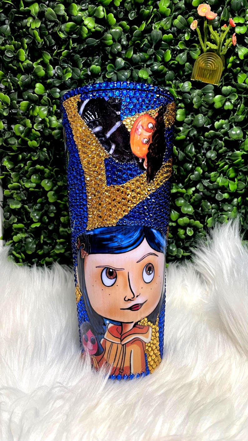 May include: A blue and gold tumbler with a rhinestone design. The tumbler features a cartoon image of a girl with blue hair and a red dress. The girl is holding a doll.