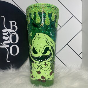 Peut inclure: Un gobelet vert &agrave; paillettes avec un personnage de dessin anim&eacute; vert et blanc avec un visage cousu et un large sourire. Le personnage porte une robe &agrave; capuche verte. Le gobelet a un couvercle vert &agrave; paillettes avec un effet de goutte &agrave; goutte.