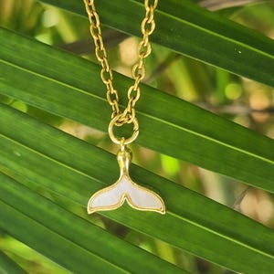 May include: A gold-toned chain necklace featuring a pendant shaped like a whale tail. The pendant has a metallic gold border and a smooth, iridescent white inlay that fills the center of the tail shape, suspended from a simple gold link chain.