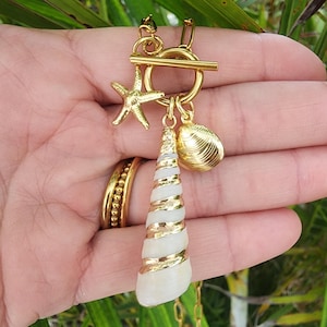 May include: A gold-toned necklace featuring a toggle clasp with three charms: a small gold starfish, a small gold clam shell, and a long, spiraled white seashell wrapped in gold metal. The necklace chain consists of elongated oval links.