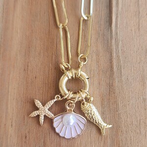 May include: A gold-toned chain necklace featuring a large circular clasp. Three charms hang from the clasp: a textured gold starfish, a light pink enamel seashell with a central white pearl bead, and a textured gold fish-shaped charm.