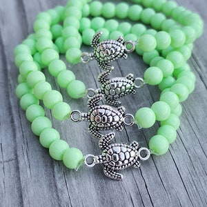 May include: Four green beaded bracelets with silver turtle charms. The bracelets are stacked on top of each other.