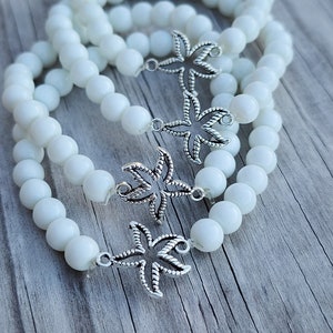 May include: Three white beaded bracelets with silver palm tree charms. The bracelets are stacked on top of each other.
