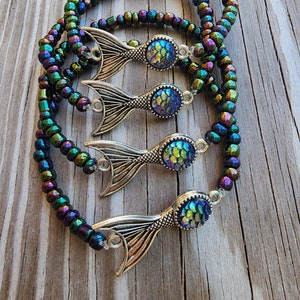 May include: Three iridescent mermaid tail charm bracelets with a dark green, purple, and blue color scheme. The charms are silver with iridescent scales.