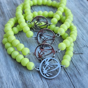 May include: Three lime green beaded bracelets with a silver palm tree charm on each. The bracelets are stacked on top of each other on a wooden surface.