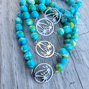 May include: Three turquoise and white patterned beaded bracelets with silver palm tree charms. The bracelets are layered on a wooden surface.