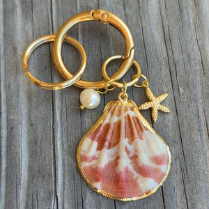 May include: A gold-toned metal keychain featuring two circular rings, a single white pearl bead, a small gold-toned starfish charm, and a large, fan-shaped seashell with white and reddish-brown radial patterns, all connected by gold-toned jump rings.