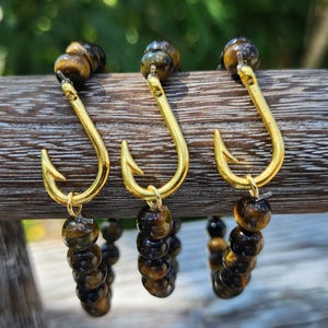 May include: Three gold fishing hook charms attached to tiger's eye beaded bracelets. The bracelets are brown and gold.