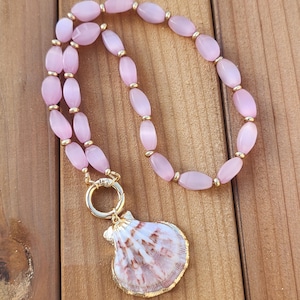 May include: A necklace featuring a series of oval-shaped, translucent light pink beads interspersed with small, round gold-toned metal spacers. The necklace includes a large gold-toned circular clasp attached to a single, natural seashell pendant.