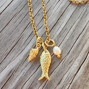 May include: A gold-toned metal necklace featuring three charms suspended from a delicate chain. The charms include a textured seashell, a detailed fish pendant with embossed scales, and a single small white freshwater pearl bead.