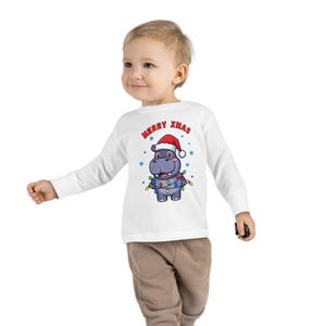 Puede incluir: Camiseta blanca de manga larga para niños pequeños con un hipopótamo de dibujos animados que lleva un gorro de Papá Noel y luces navideñas. La camiseta tiene el texto "MERRY XMAS" en rojo. El hipopótamo es gris y azul, con copos de nieve azules alrededor.