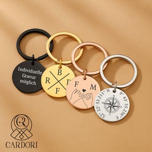 Könnte beinhalten: Eine Auswahl an personalisierten Schlüsselanhängern mit verschiedenen Designs. Ein schwarzer Schlüsselanhänger trägt den Text "Individuelle Gravur möglich". Weitere Schlüsselanhänger zeigen Initialen, verschränkte Hände mit Herz und einen Kompass mit Koordinaten.