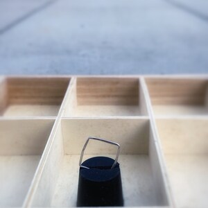 May include: A small, silver-colored ring with a square top is displayed on a black velvet stand inside a light-colored wooden organizer. The organizer has multiple square compartments.