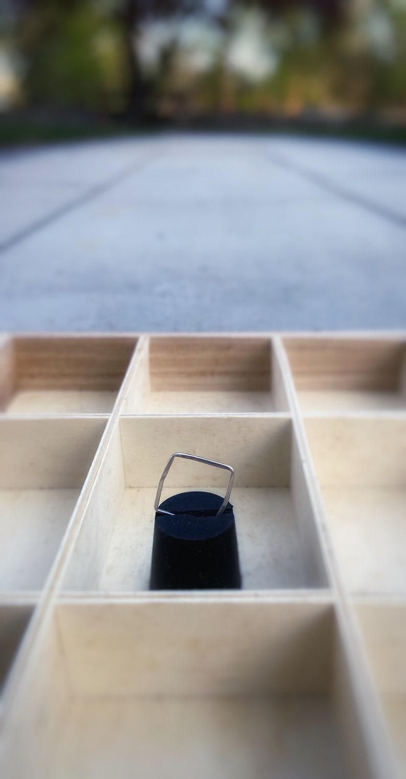 May include: A small, silver-colored ring with a square top is displayed on a black velvet stand inside a light-colored wooden organizer. The organizer has multiple square compartments.