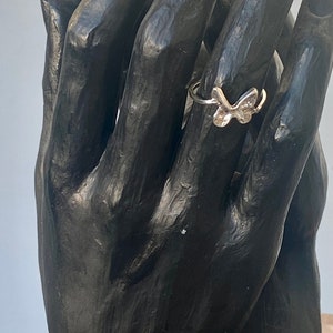 May include: A silver butterfly ring. The ring features a delicate butterfly design with detailed wings. The ring is displayed on a black sculpture of hands, showcasing the ring's design and size.