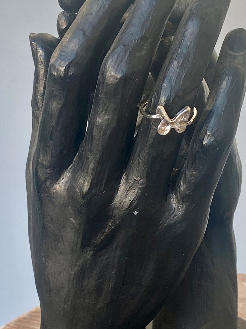 May include: A silver butterfly ring. The ring features a delicate butterfly design with detailed wings. The ring is displayed on a black sculpture of hands, showcasing the ring's design and size.