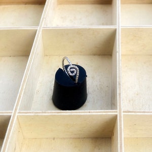 May include: A silver ring with a spiral design, set on a black cylindrical object. The ring is displayed in a light-colored wooden box with multiple compartments. The ring's spiral is made of small, sparkling stones.