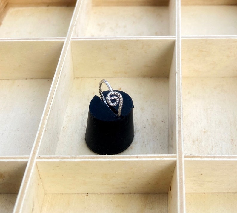 May include: A silver ring with a spiral design, set on a black cylindrical object. The ring is displayed in a light-colored wooden box with multiple compartments. The ring's spiral is made of small, sparkling stones.