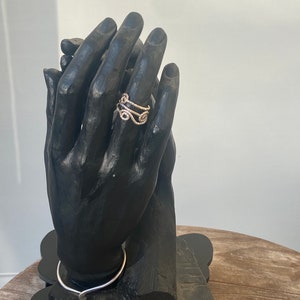 May include: A gold-colored ring with an intricate, swirling design is displayed on a black hand-shaped jewelry holder. The ring's design features multiple curves and loops. A silver-colored necklace is also visible.