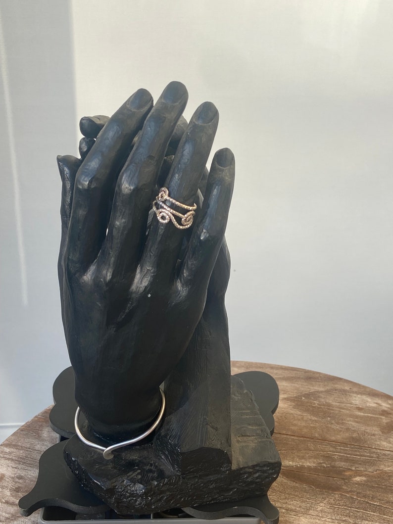 May include: A gold-colored ring with an intricate, swirling design is displayed on a black hand-shaped jewelry holder. The ring's design features multiple curves and loops. A silver-colored necklace is also visible.