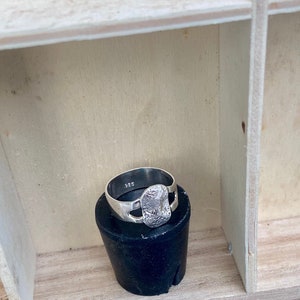 May include: A silver ring with a textured, rectangular center stone. The ring is marked with "925" on the inside of the band. The ring is displayed on a black cylindrical object, set against a light wooden background.