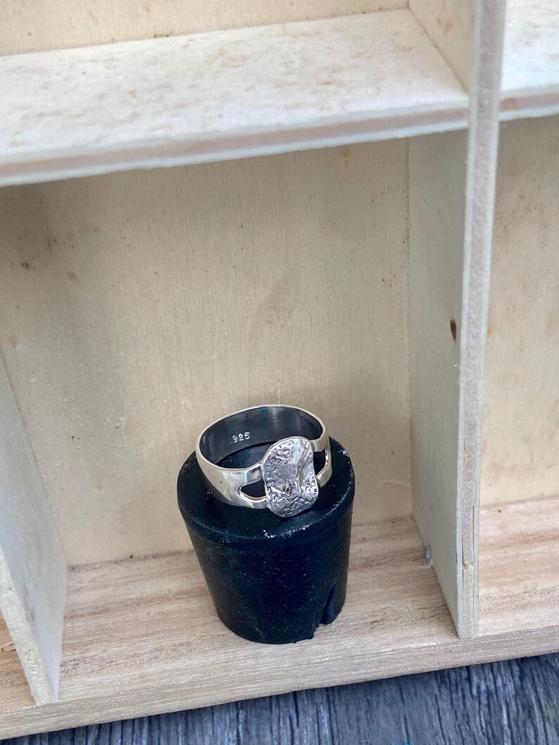 May include: A silver ring with a textured, rectangular center stone. The ring is marked with "925" on the inside of the band. The ring is displayed on a black cylindrical object, set against a light wooden background.
