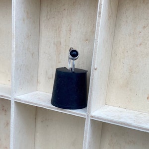 May include: A silver ring with a round, black gemstone. The ring is displayed on a black, conical object, set against a backdrop of a light-colored, divided wooden shelf.