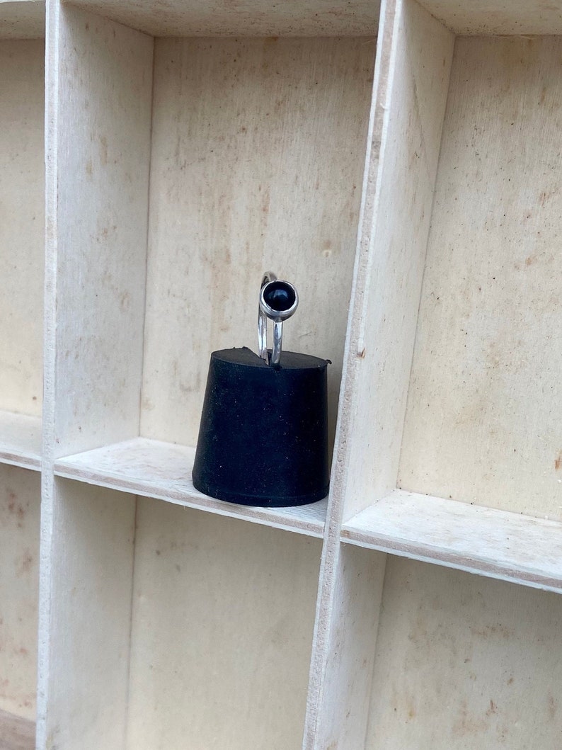 May include: A silver ring with a round, black gemstone. The ring is displayed on a black, conical object, set against a backdrop of a light-colored, divided wooden shelf.