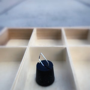 May include: A black rubber stopper with a silver metal handle, set inside a light-colored wooden organizer with multiple compartments. The stopper is the focal point, with the handle angled upwards.