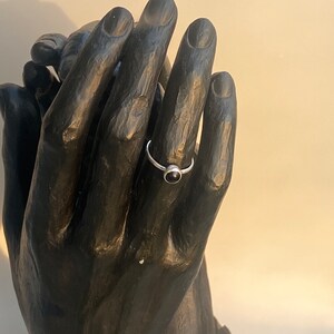 May include: A silver ring with a small, round, black gemstone. The ring is displayed on a black hand sculpture. The ring's band is thin and appears to be made of silver. The gemstone is set in a simple bezel setting.