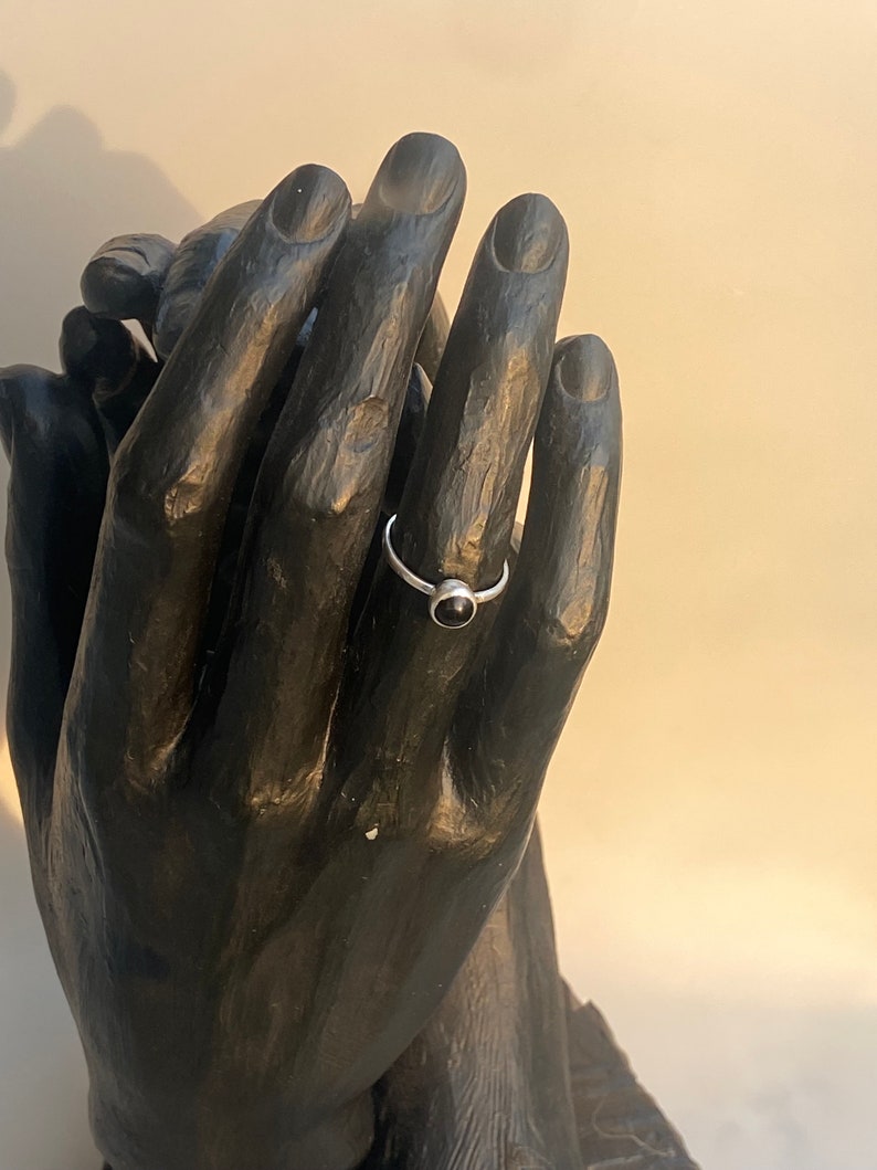 May include: A silver ring with a small, round, black gemstone. The ring is displayed on a black hand sculpture. The ring's band is thin and appears to be made of silver. The gemstone is set in a simple bezel setting.