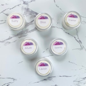 May include: Six small clear jars with white lids, each containing a white cream. Each jar has a white label with a purple design and a different word printed in black: Sultry, Bliss, Enarmor, Allure, Serenity, Lavish.