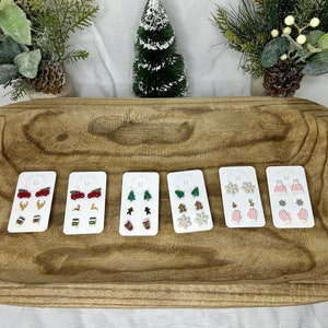 May include: Six sets of Christmas-themed earrings on white cards. Each set includes three pairs of earrings with different designs, including Christmas trees, reindeer, snowflakes, mittens, coffee cups, and pickup trucks.