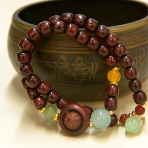 May include: A dark red wooden beaded bracelet with green and orange beads, and a large dark red wooden bead in the center. The bracelet is resting on a dark brown bowl with intricate designs.