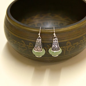 May include: Silver-tone drop earrings featuring a light green, teardrop-shaped stone. The earrings have an ornate, filigree design at the top. The earrings are displayed in front of a dark, patterned bowl.