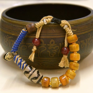 Puede incluir: Un brazalete de cuentas multicolores con un cordón azul, blanco y marrón. El brazalete presenta cuentas de color ámbar, cuentas de madera marrón y una cuenta con un patrón en blanco y negro. El brazalete está apoyado en un cuenco de madera marrón con intrincados tallados.