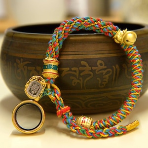 May include: A colorful braided bracelet with red, blue, green, and gold threads. The bracelet features gold-colored metal accents, including a small bell, decorative beads, and a pendant with a face design. The bracelet is displayed near a bronze bowl.