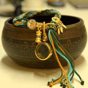 Pode incluir: Um bracelete de ouro com vários pingentes, incluindo uma roda de oração, uma pequena esfera de vidro e um pingente de flor colorido. O bracelete é feito de cordões trançados em tons de verde, laranja e turquesa.