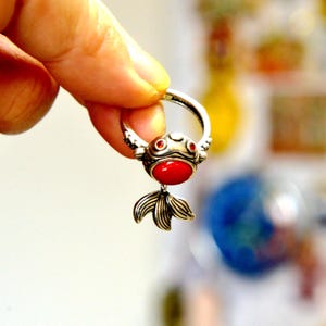 May include: A silver ring featuring a goldfish design. The ring has a red oval-shaped stone for the fish's mouth and small red stones for the eyes. The fish's tail is a detailed, stylized fan shape.
