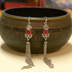 May include: A pair of silver-toned dangle earrings with a rectangular red stone set in an ornate frame. The earrings have a chain tassel design. The background is a bronze-colored bowl with etched patterns.