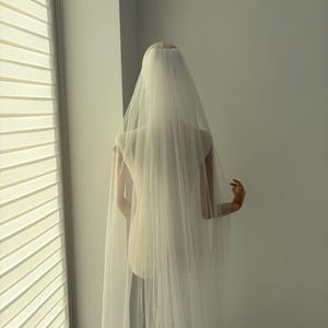 May include: A sheer, ivory-colored bridal veil cascading down the back of a mannequin. The veil is long and flowing, with a delicate texture. The mannequin is positioned in front of a window with blinds.