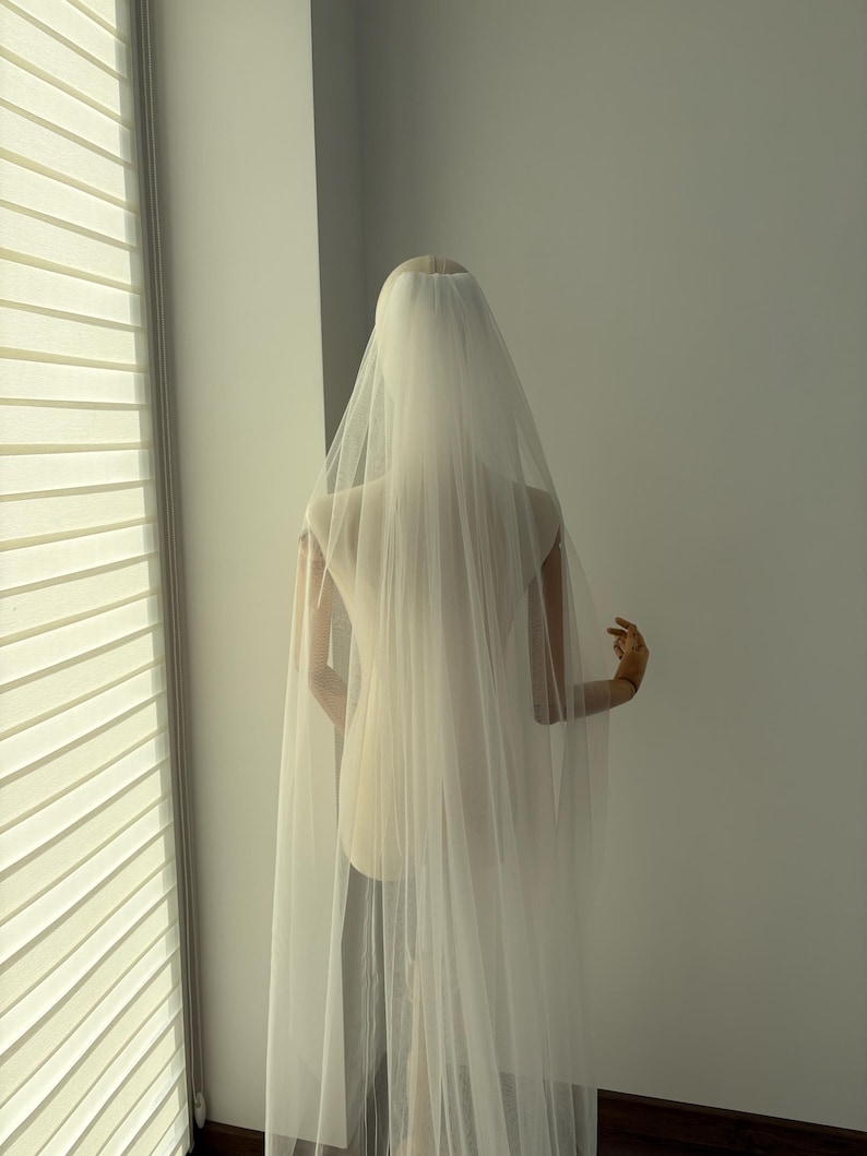 May include: A sheer, ivory-colored bridal veil cascading down the back of a mannequin. The veil is long and flowing, with a delicate texture. The mannequin is positioned in front of a window with blinds.