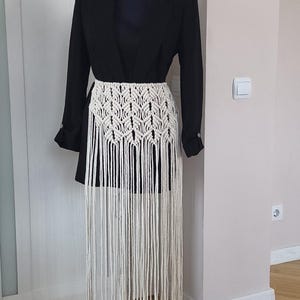 May include: A cream macrame skirt with a geometric design and long fringe is displayed on a black blazer. The skirt's intricate design and flowing fringe create a bohemian aesthetic. The blazer is on a mannequin.